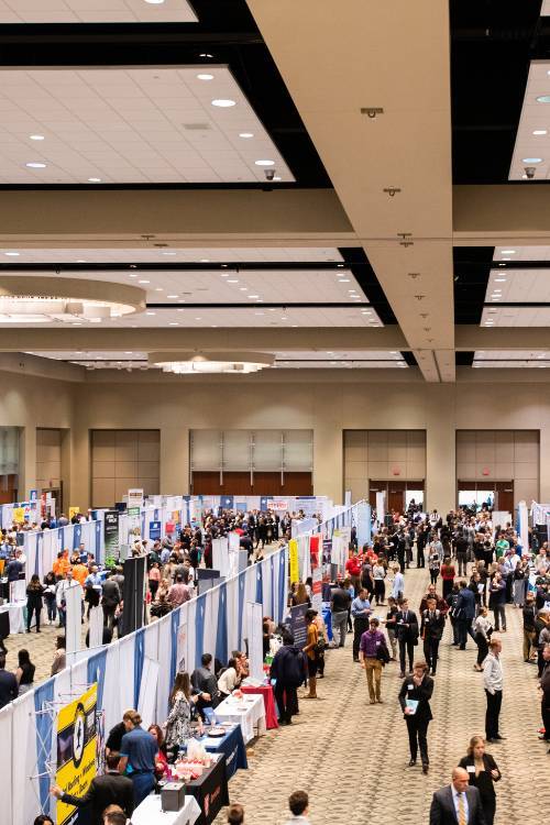 Large room with booths in multiple rows with many people walking down the aisles